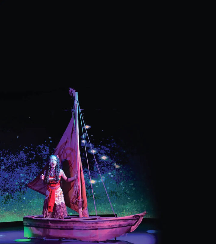 A performer stands in a lit sailboat on a dark stage